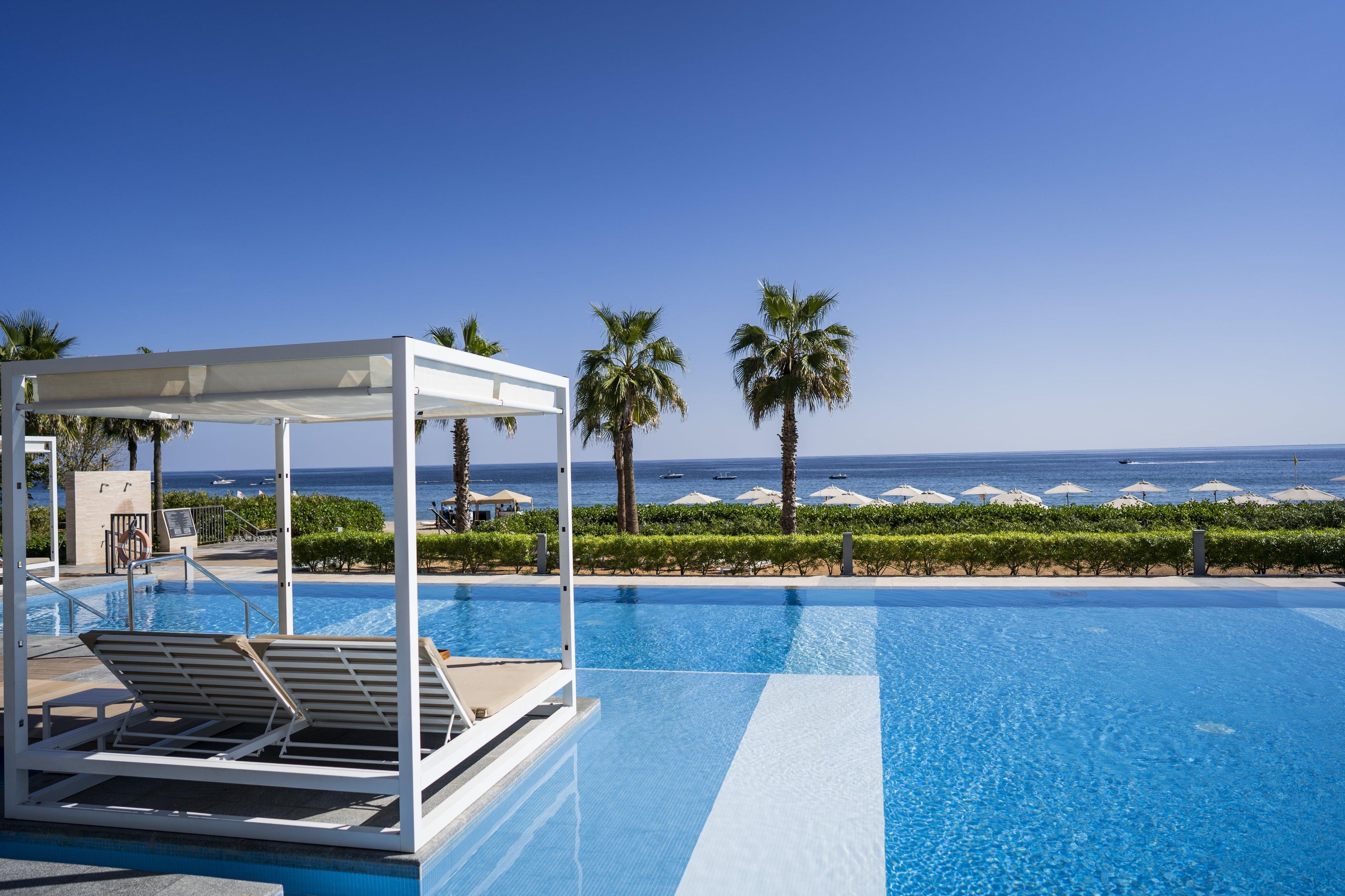 A clear blue pool with lounge chairs, palm trees, and a view of the ocean.