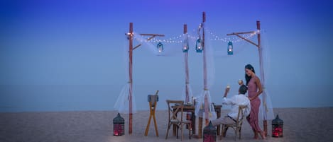 Escena de playa con una estructura de madera, faroles y una mesa con sillas.