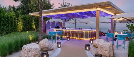 A beachside bar with a wooden deck, seating, and a view of the ocean.