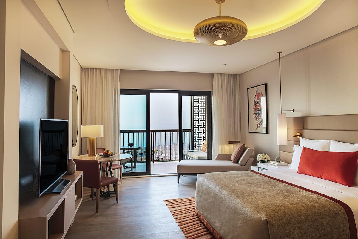 A hotel room with a bed, a TV, a desk with a chair, a balcony with a view, and a large window.