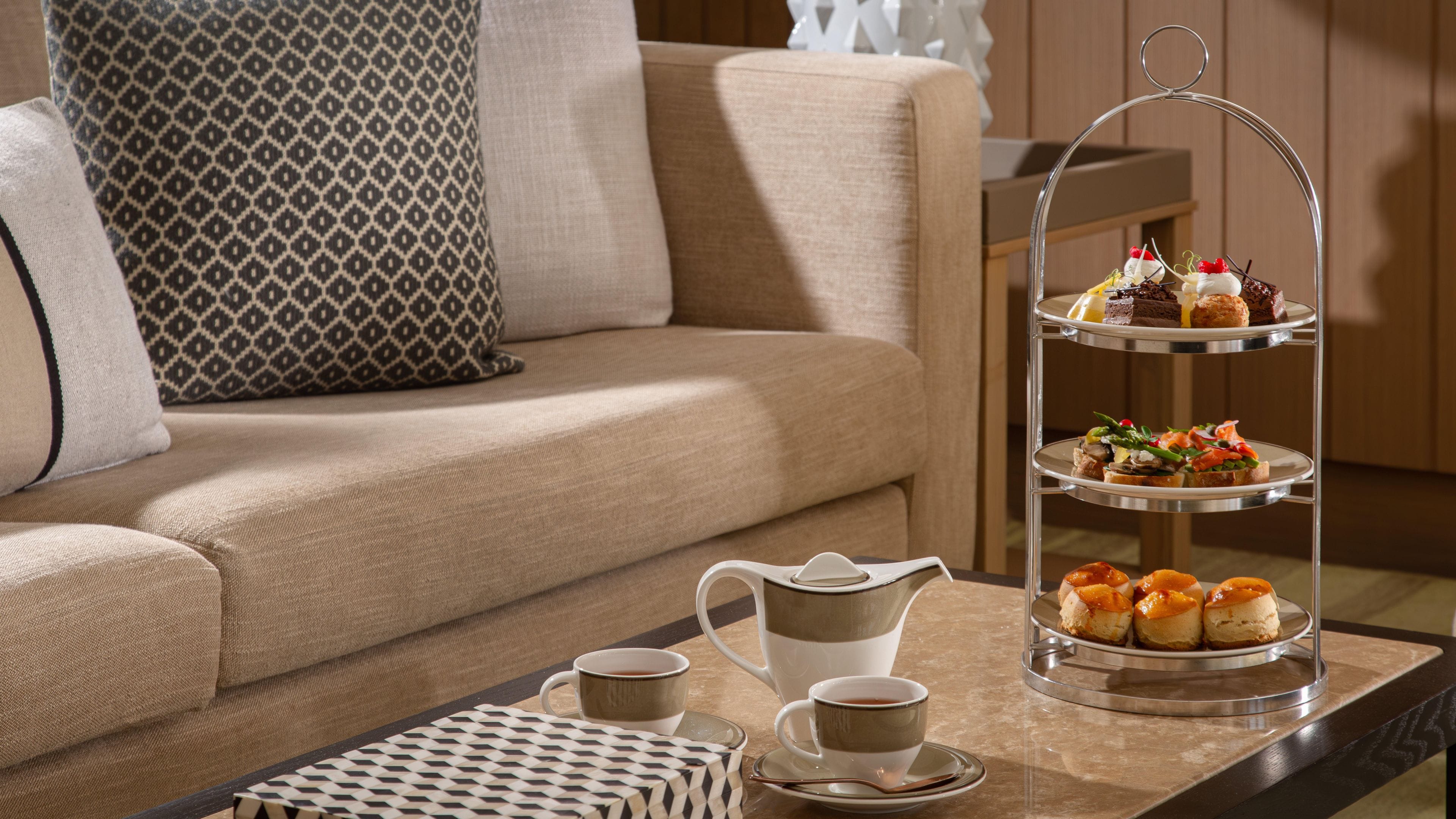 A three-tiered dessert stand with various pastries, a coffee pot, cups, and a tray with a patterned box on a table in a room with a sofa and patterned pillows.