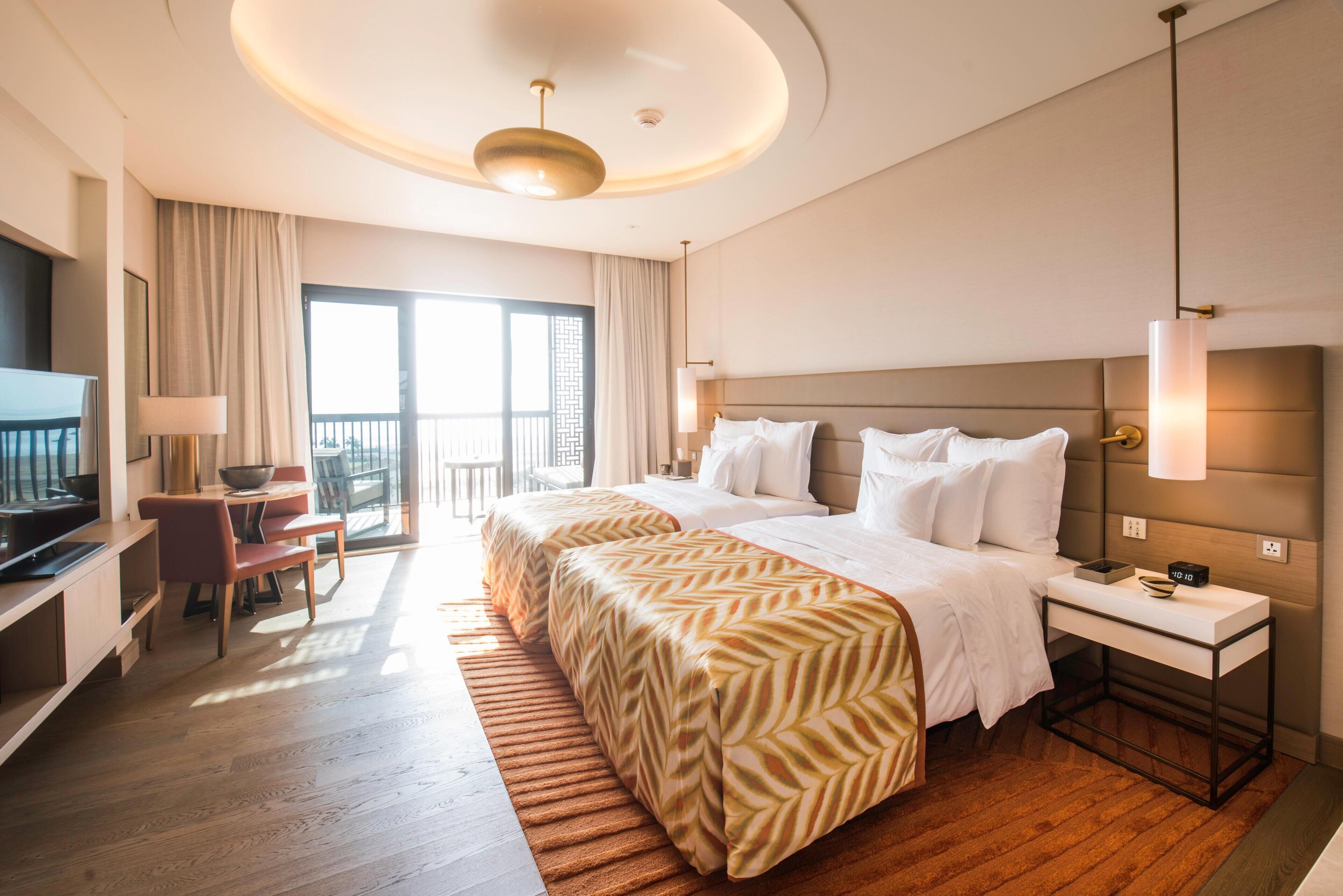 A hotel room with a bed, bedside table, lamp, TV, desk, chair, and a balcony.