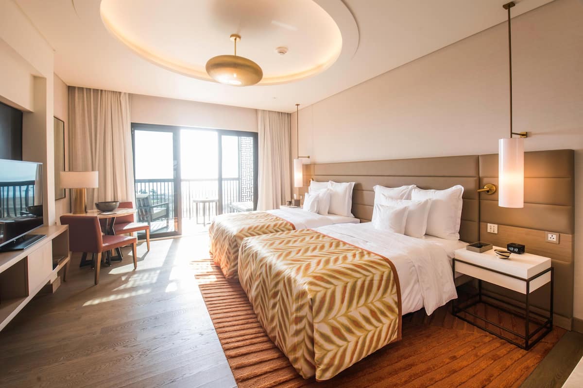 A hotel room with a bed, bedside table, lamp, TV, desk, chair, and a balcony.