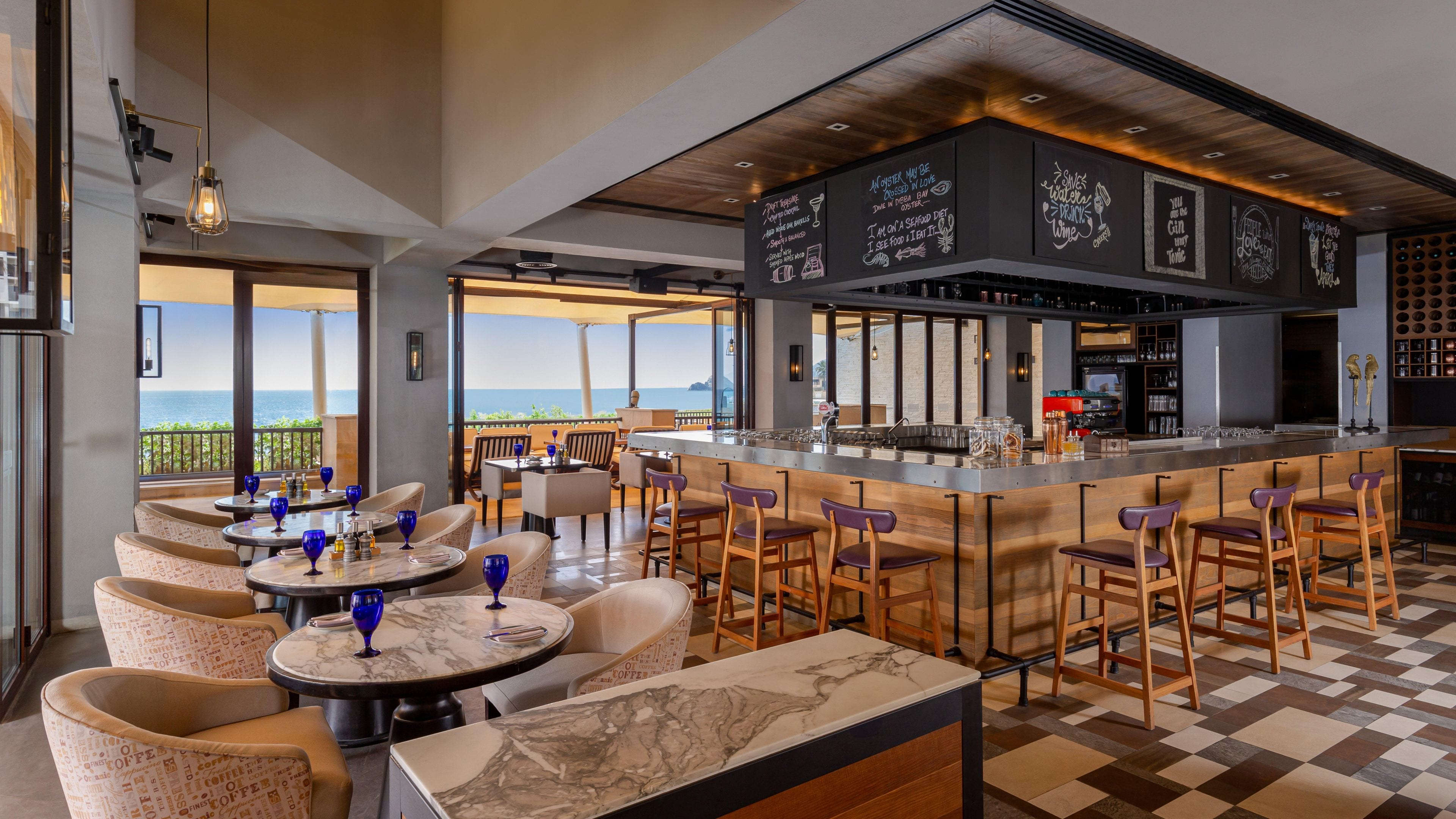 A bar area with a view of the sea, seating with a checkered floor, and a chalkboard menu.