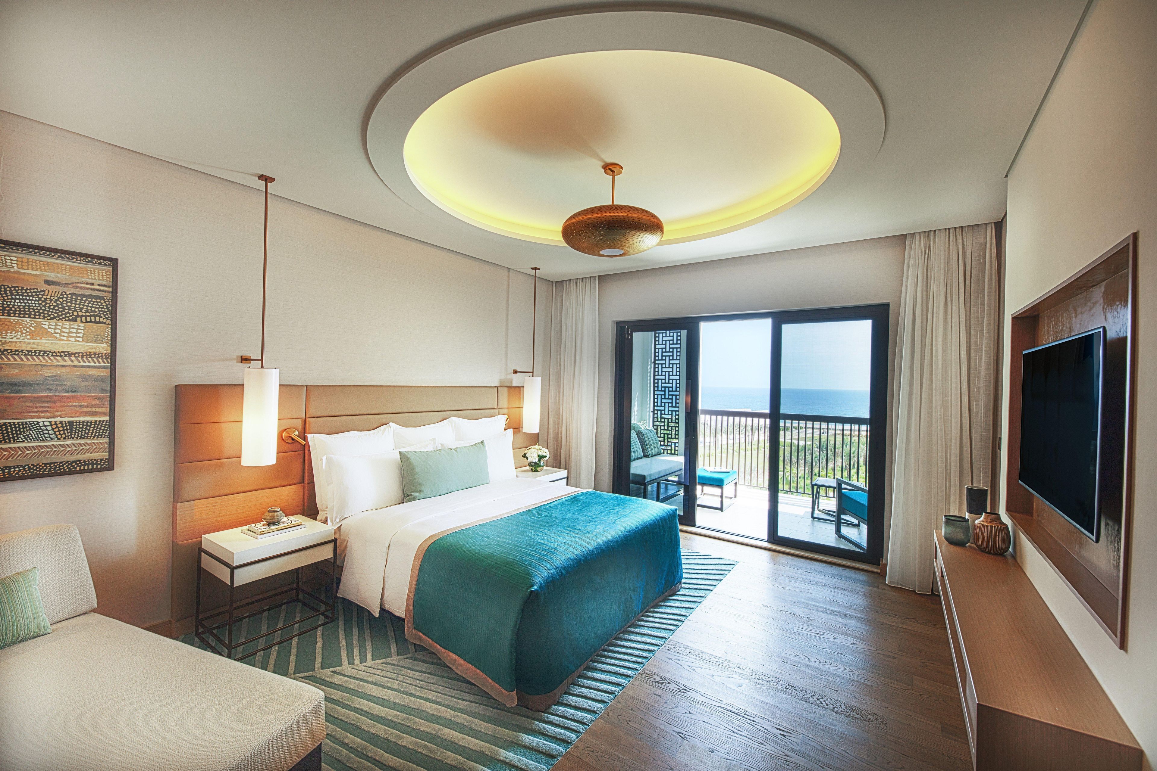 A hotel room with a large bed, a desk, a chair, a TV, and a balcony with a view.