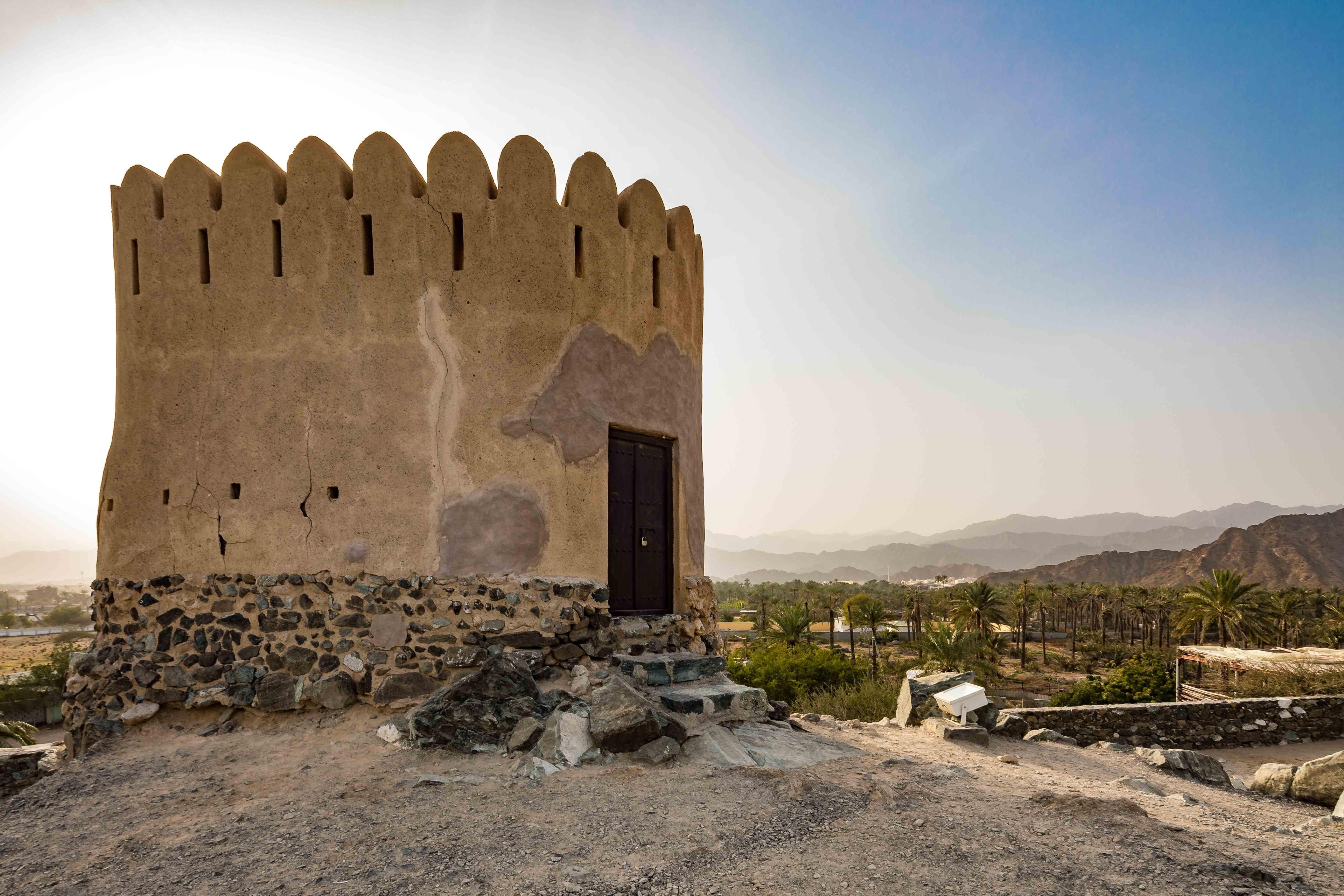 A stone tower with a dark door, surrounded by a rocky landscape and palm trees.