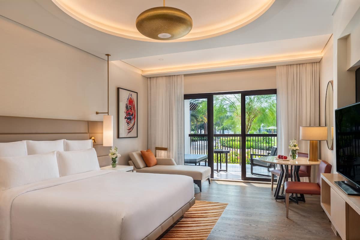 A hotel room with a large bed, a TV, a desk with a lamp, and a balcony with a view of greenery.