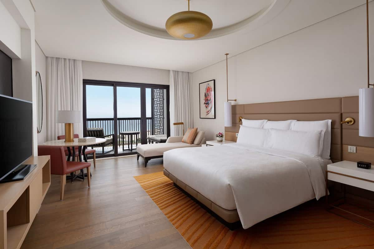 A hotel room with a large bed, a desk with a chair, a TV, a balcony with a view, and a dining area with a table and chairs.