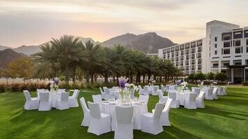 An outdoor event space with round tables and white chairs, set against a backdrop of palm trees and mountains, with a modern building in the background.