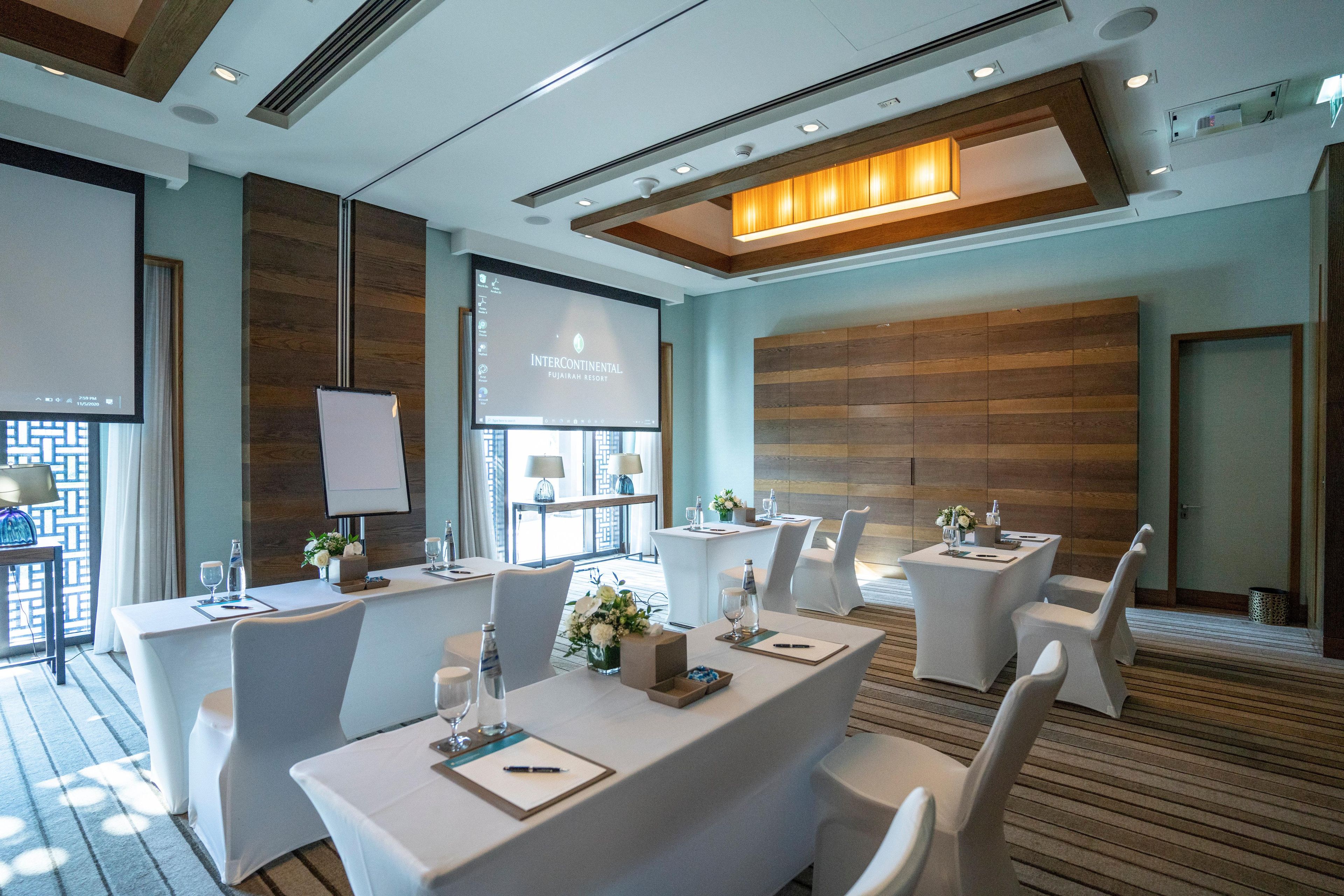 A conference room with white tables, chairs, a projector screen, and a wooden panel wall.