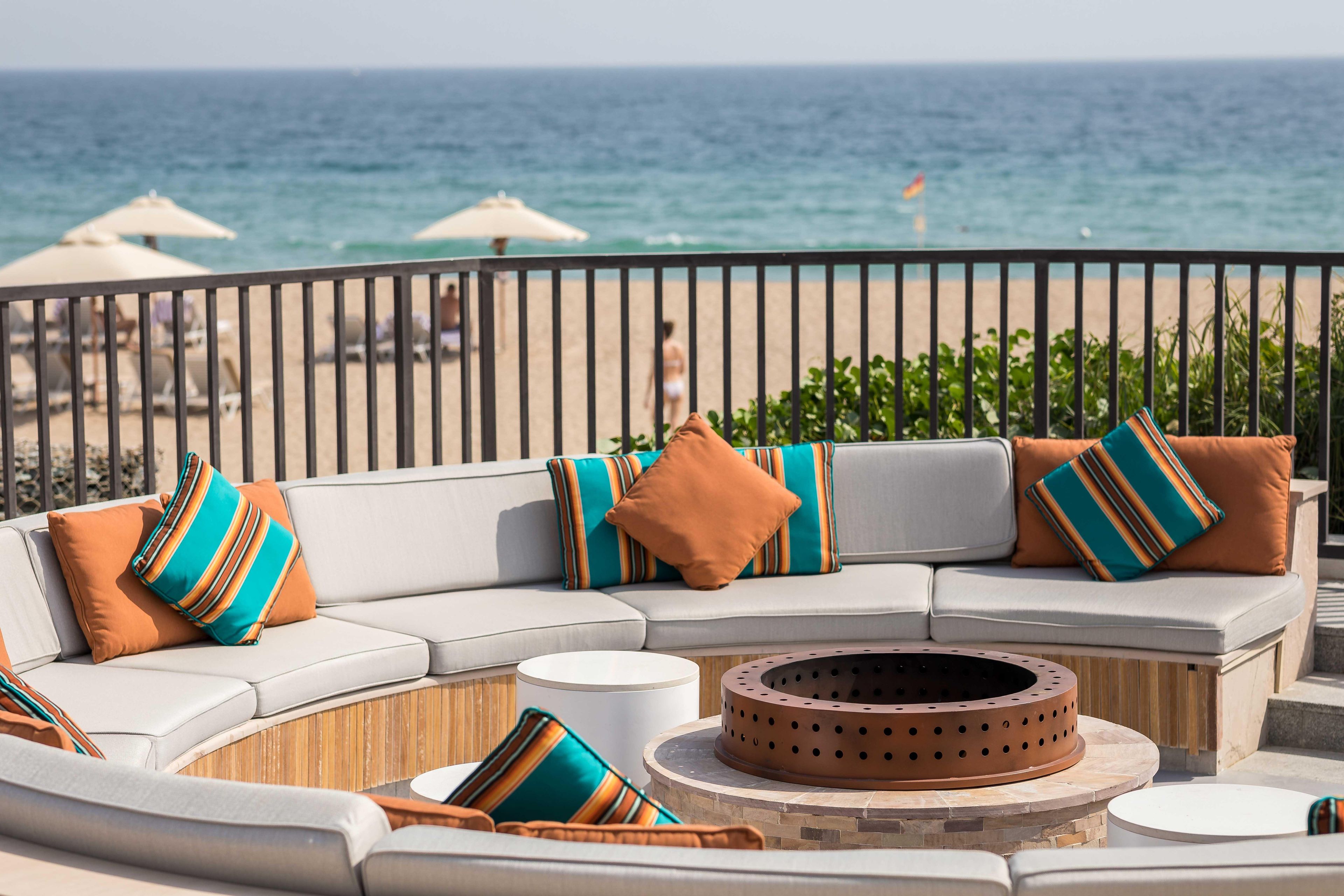 A curved outdoor seating area with a fire pit, white round tables, and colorful cushions.