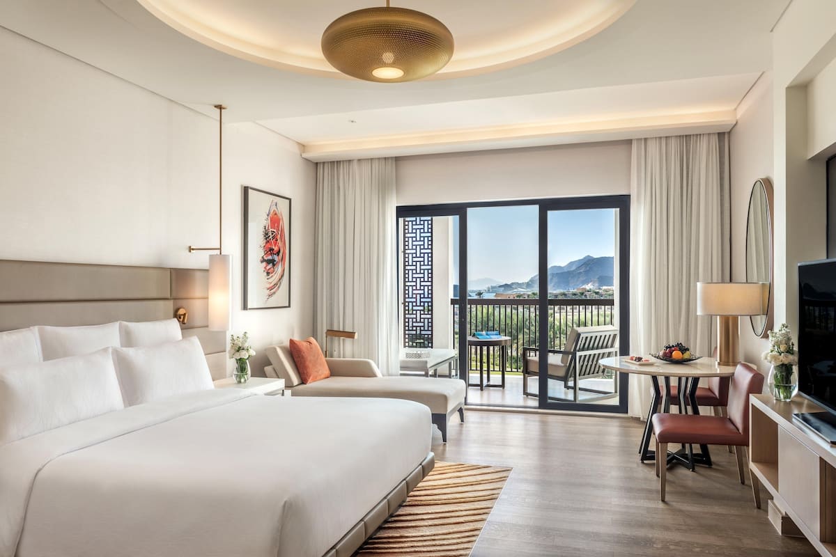 A hotel room with a large bed, a desk with a chair, a television, and a balcony with a view.