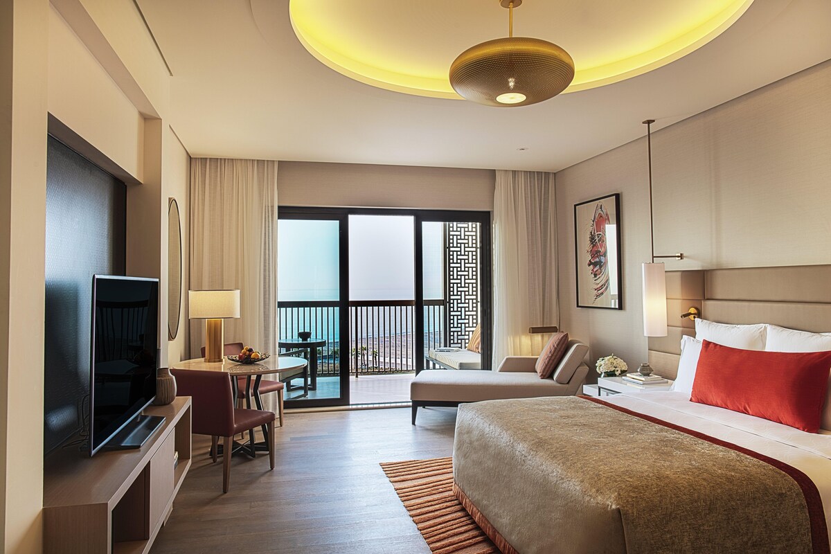 A hotel room with a bed, a TV, a desk with a chair, a balcony with a view, and a large window.