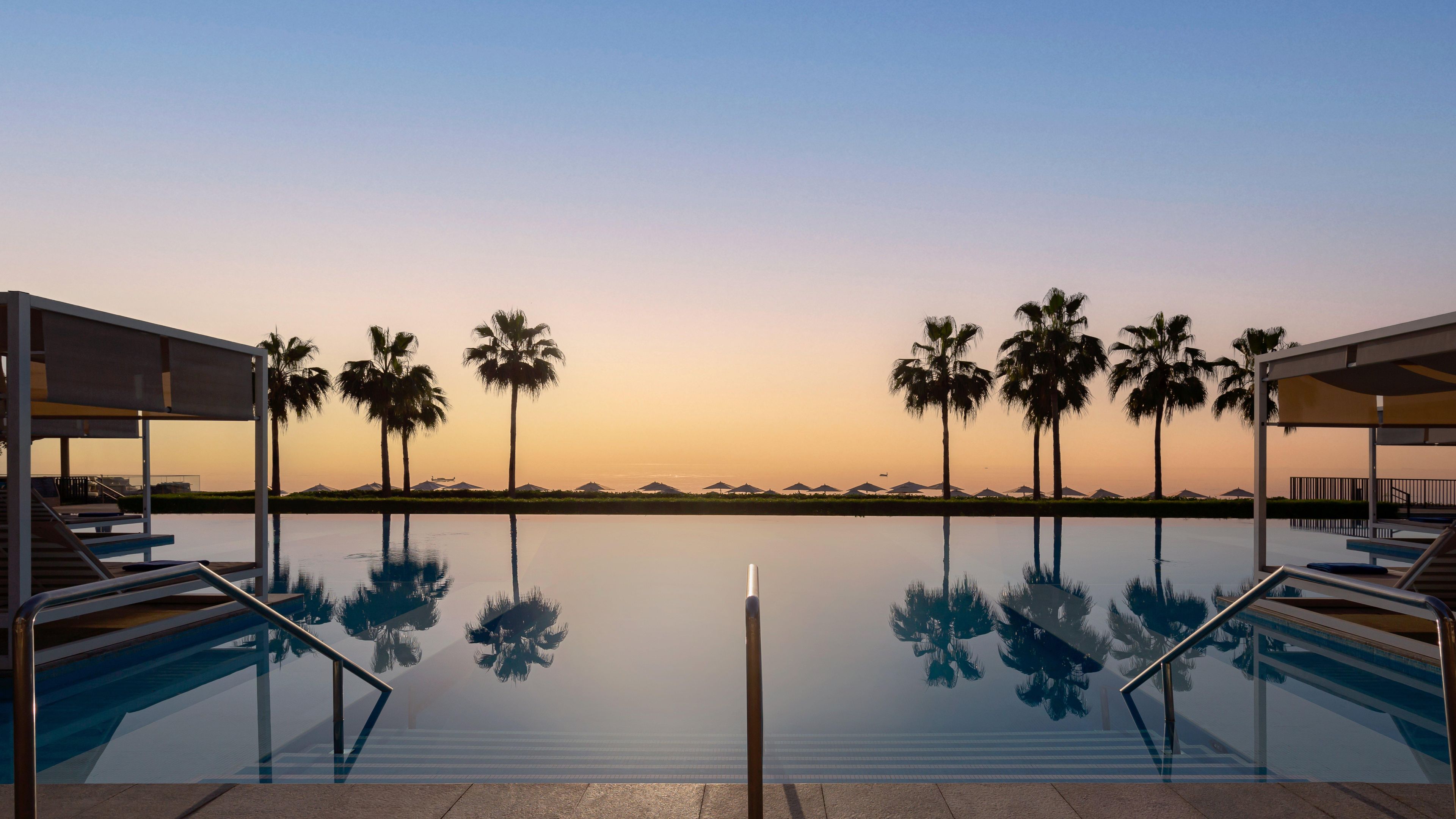 Una piscina con agua cristalina que refleja palmeras y un atardecer al fondo.