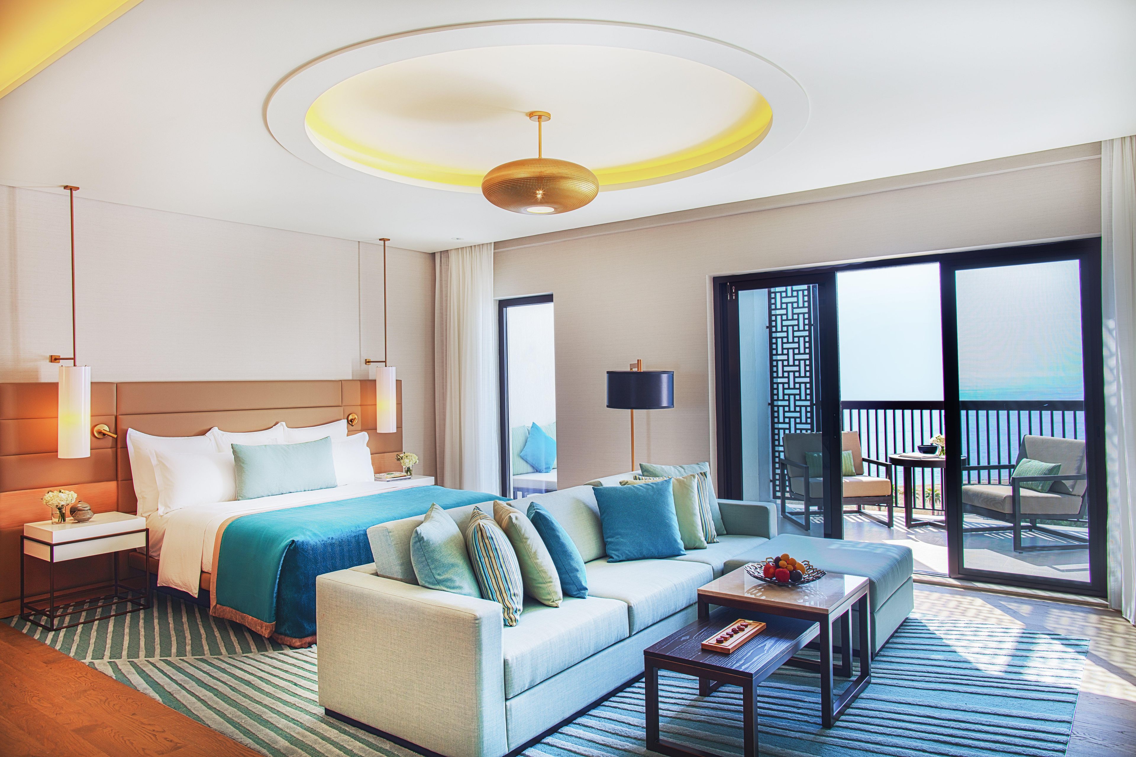A hotel room with a bed, a sofa, a coffee table, a nightstand, a lamp, and a view of the sea through a balcony door.