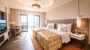 Habitación de hotel con cama, mesita de noche, lámpara, televisor, escritorio, silla y balcón.