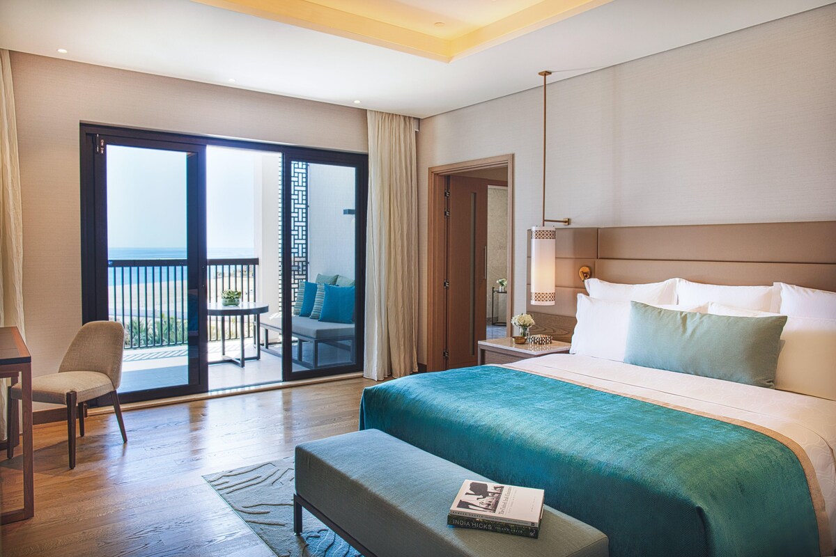 A hotel room with a large bed, a desk, a chair, a balcony with a view, and a door leading to another room.