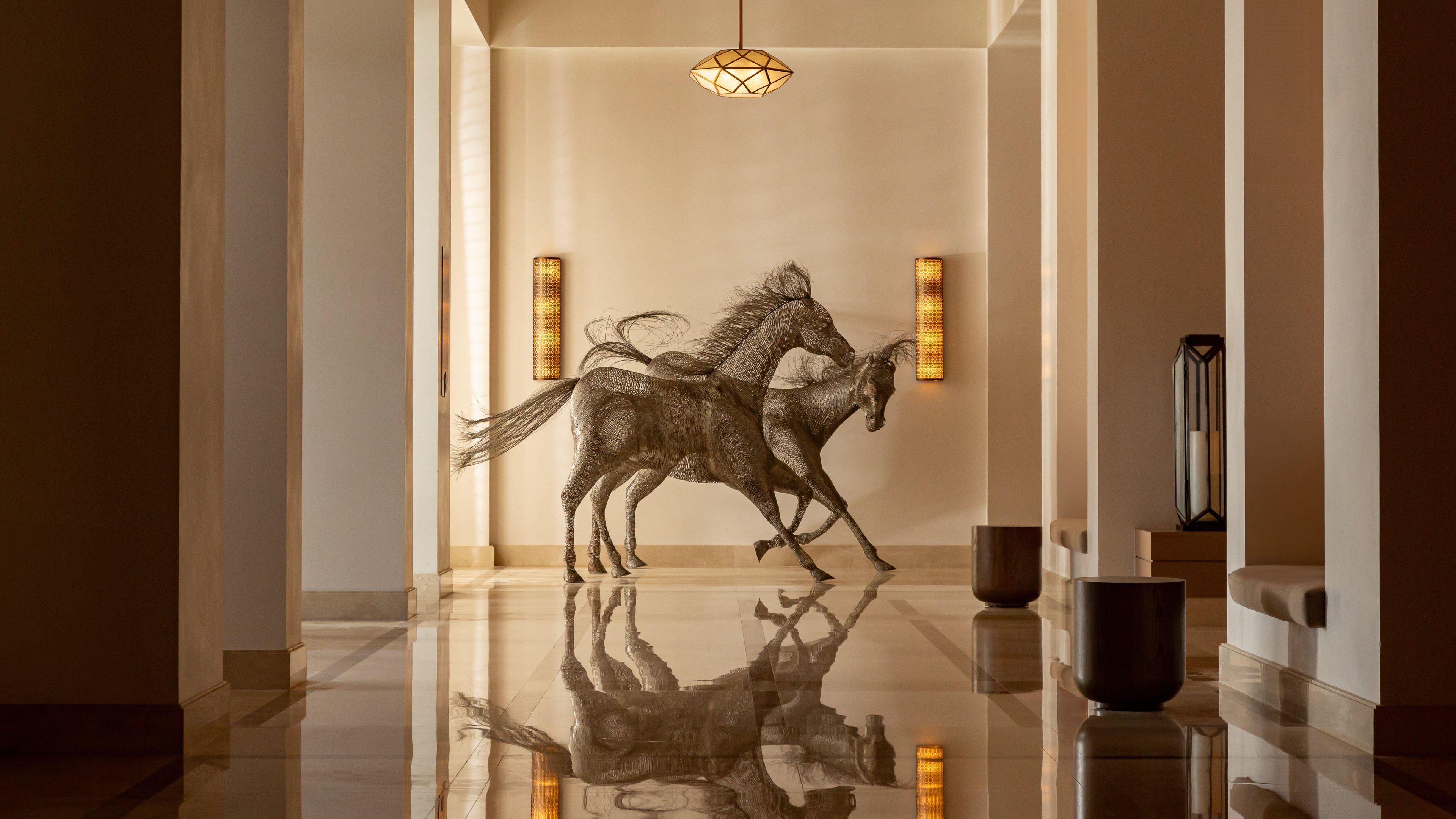 A hallway with a reflective floor, a geometric light fixture, and a wall-mounted sculpture of two horses.