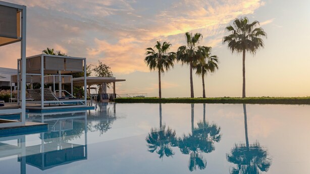 Piscina con agua cristalina que refleja palmeras y un edificio moderno con zona de estar cubierta.