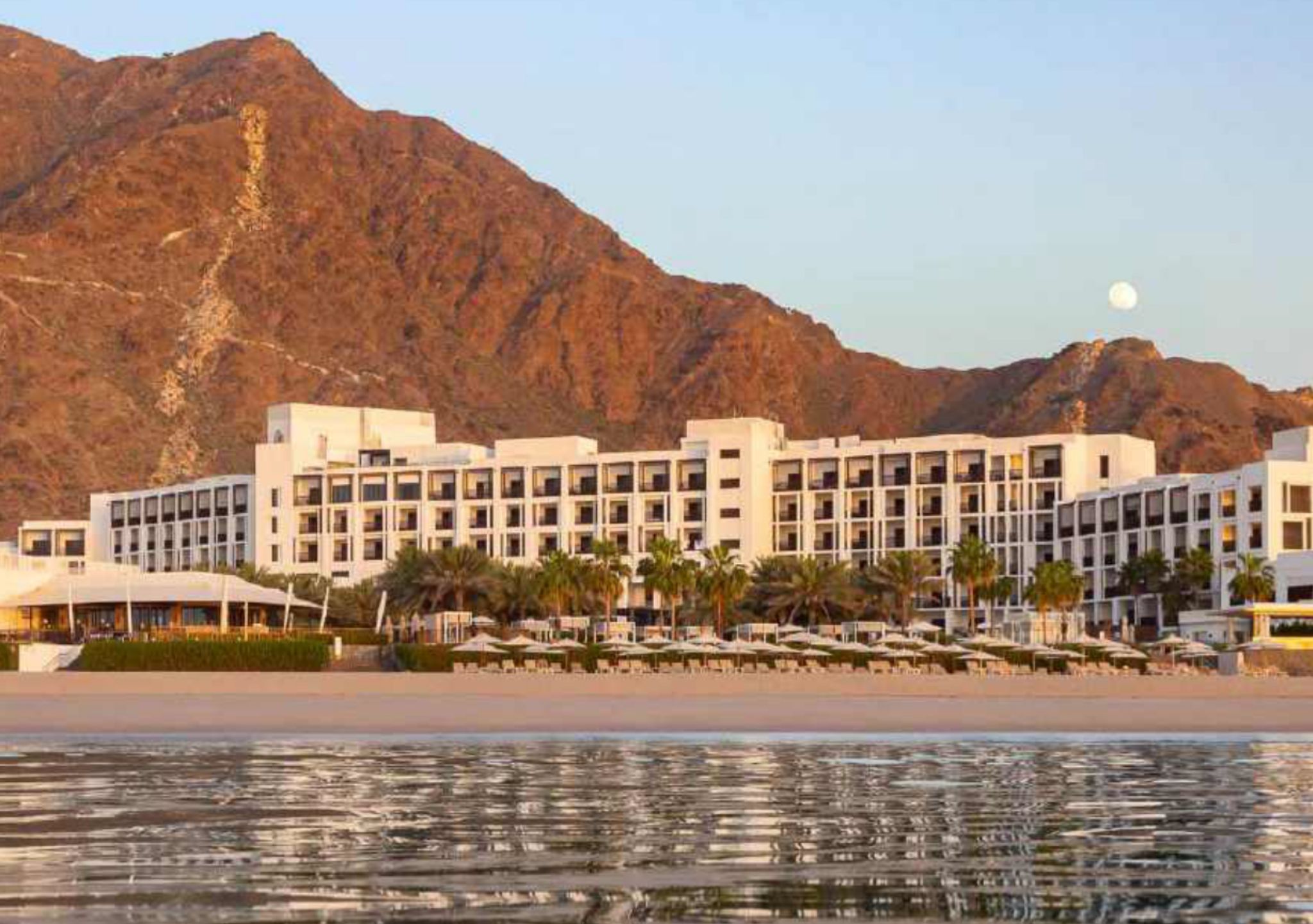 A hotel with multiple floors, a beach in the foreground, and mountains in the background.