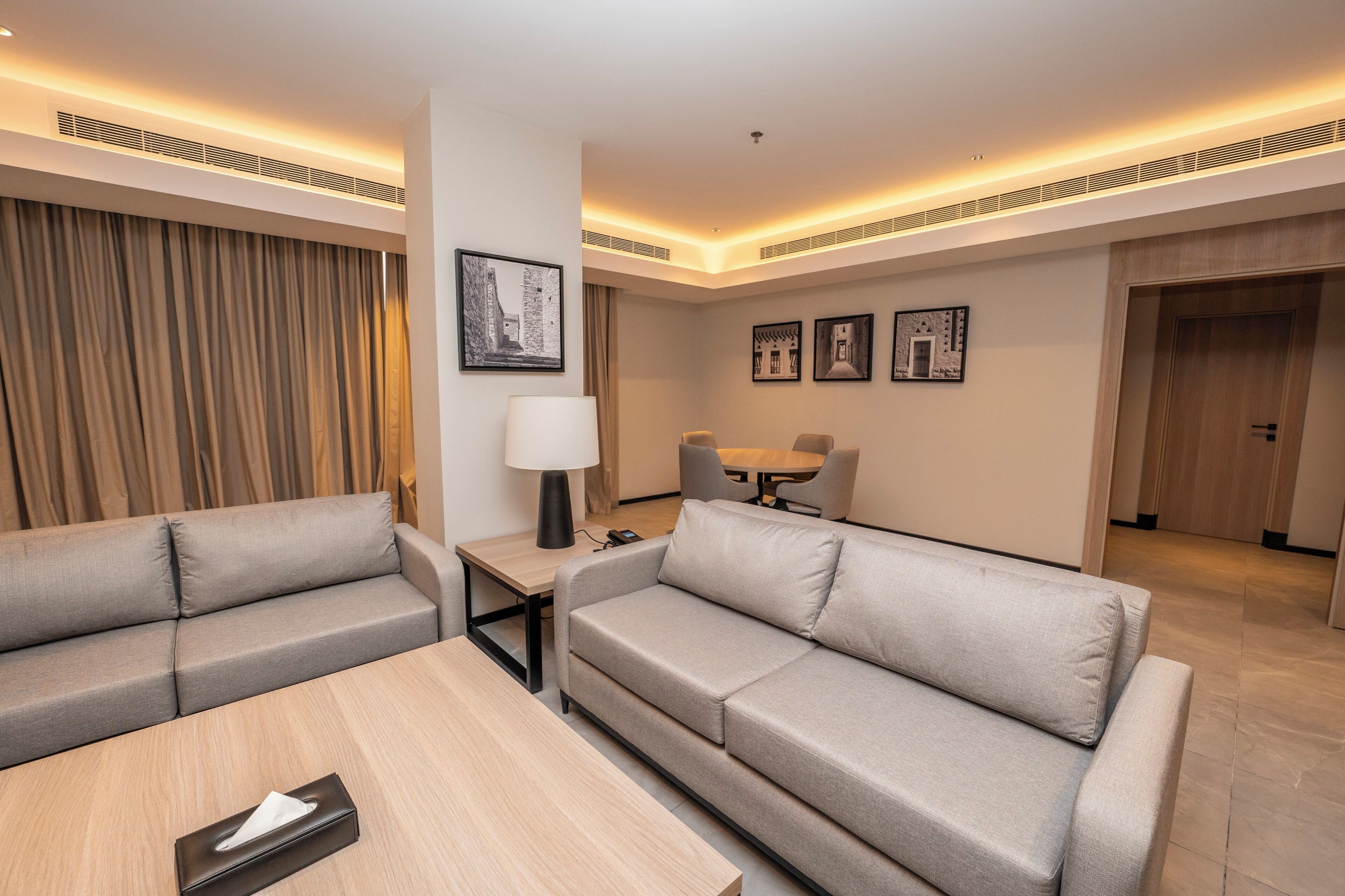 A modern hotel room with a sofa, a small table, a lamp, and a dining area with chairs.