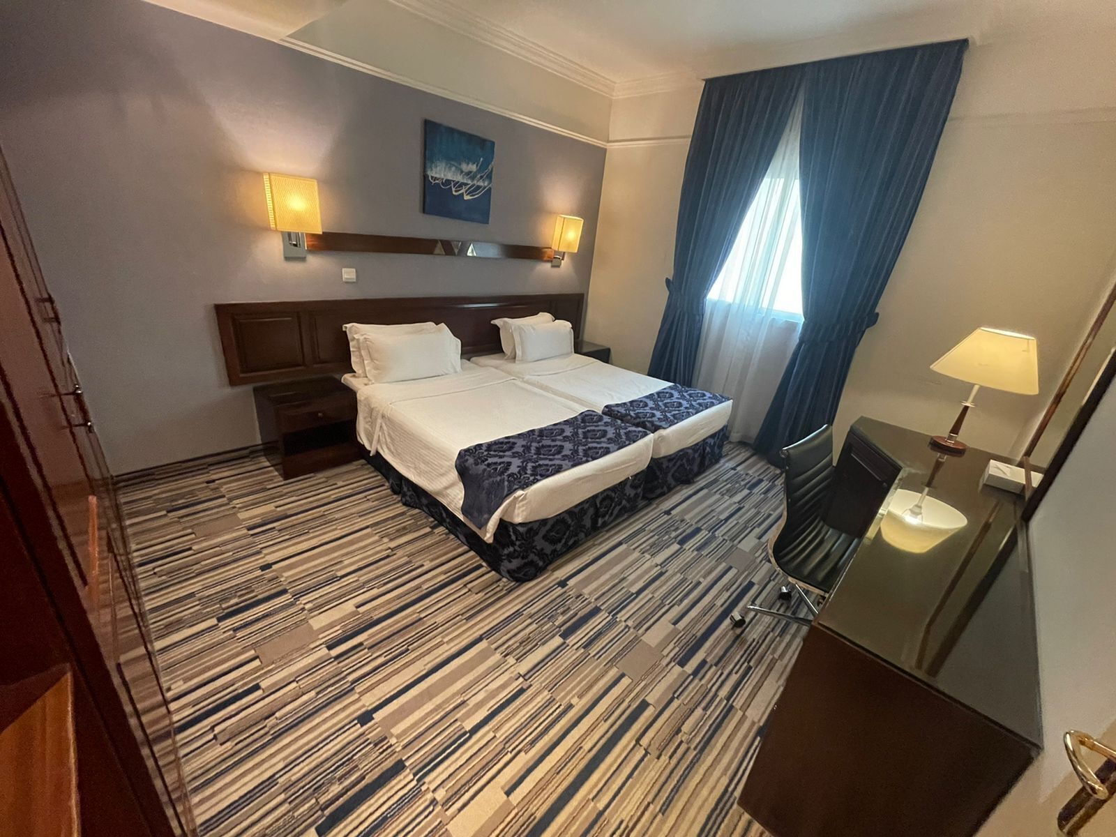 A hotel room with a large bed, two bedside tables, a desk with a chair, a mirror, a window with curtains, and a wall-mounted artwork.