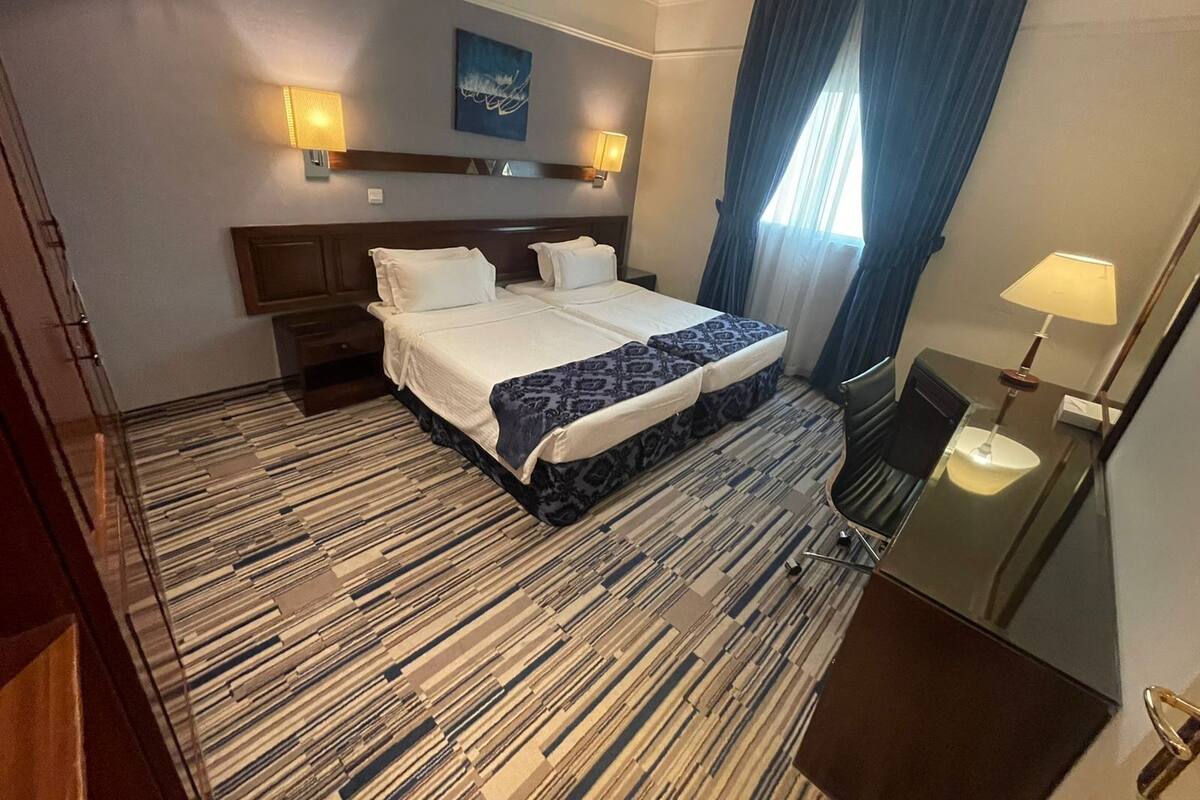 Une chambre d’hôtel avec un grand lit, deux tables de chevet, un bureau avec une chaise, un miroir, une fenêtre avec des rideaux et une œuvre d’art accrochée au mur.