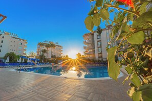 Una piscina con rayos de sol, rodeada de edificios y sillas de descanso.
