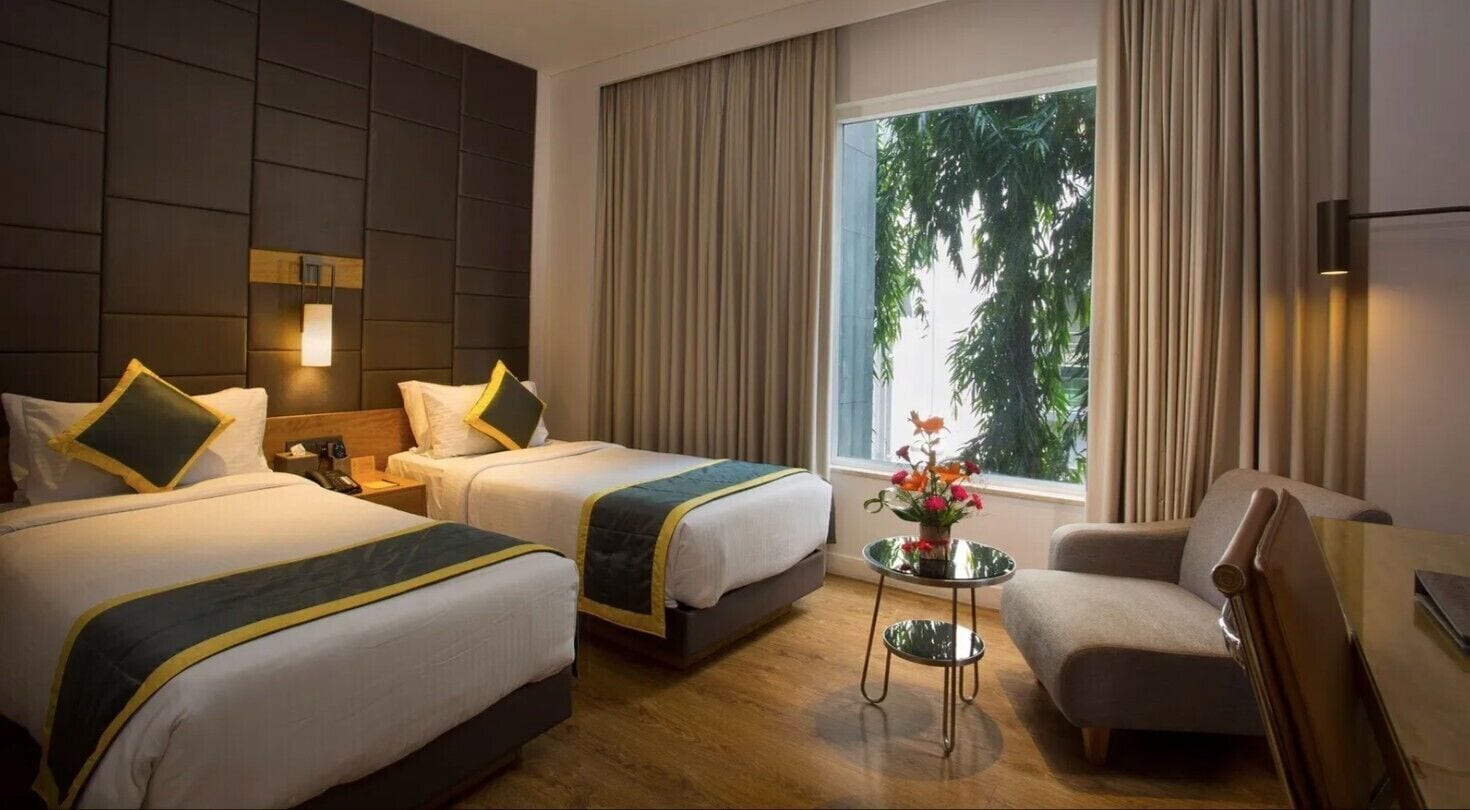 A hotel room with two beds, a small table with flowers, a chair, and a window with curtains.