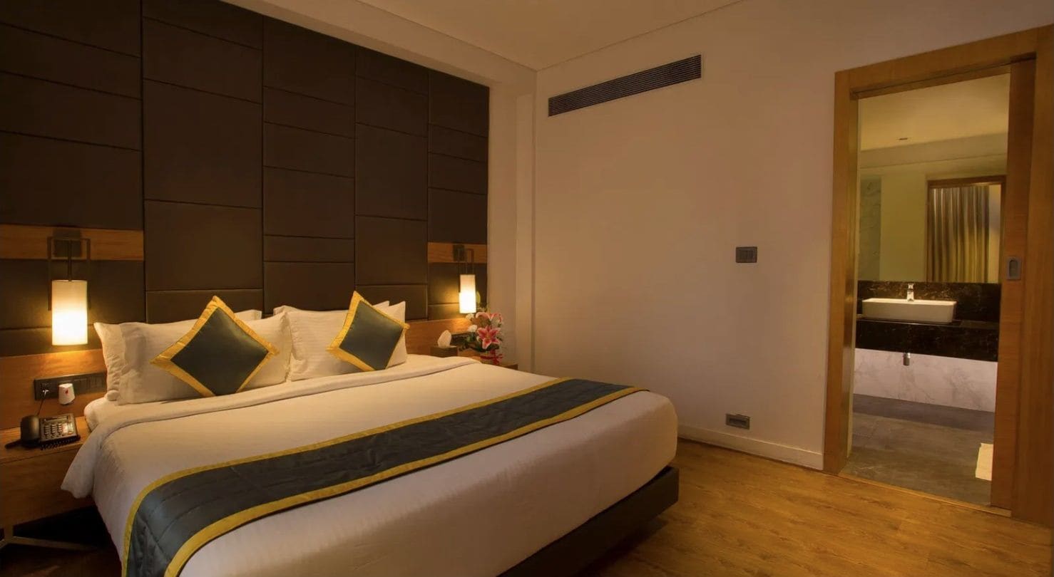 A hotel room with a large bed, bedside lamps, a wooden headboard, and a bathroom visible through an open door.