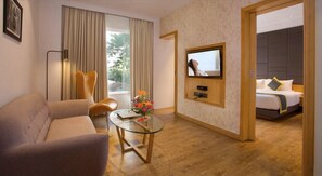 A hotel room with a sofa, a glass table with flowers, a TV, and a bed with a headboard. - Fountain Tree by TGI - JP Nagar, Bangalore (Bengaluru)