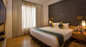 Une chambre d’hôtel avec un grand lit, une tête de lit ornée d’un motif géométrique, une table de chevet avec une lampe et une fenêtre avec des rideaux.