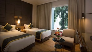 Habitación de hotel con dos camas, un sofá, una mesita con un florero y un ventanal con cortinas.