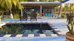 A swimming pool with tiled edges, two lounge chairs, a covered seating area with colorful chairs, and a small building with a corrugated metal roof. - Cabaña Olas Del Mar (San Bernardo del Viento)