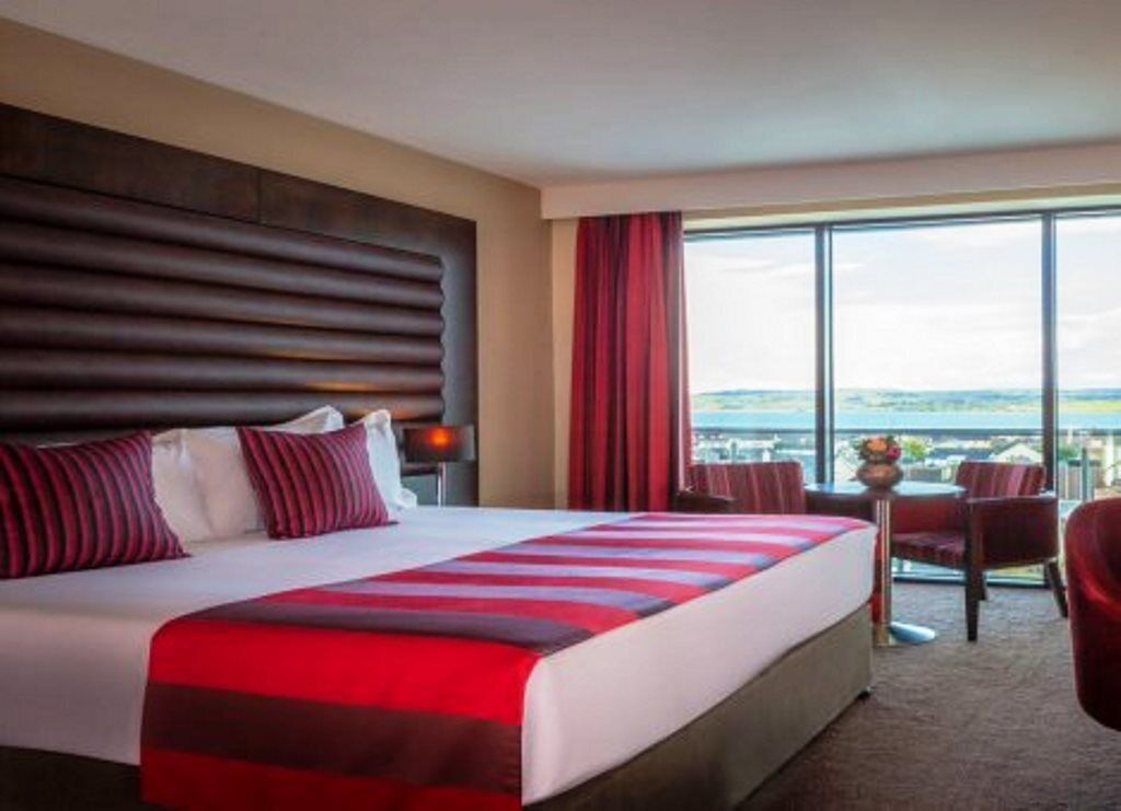 A hotel room with a large bed, a view of the city, and a balcony with chairs.
