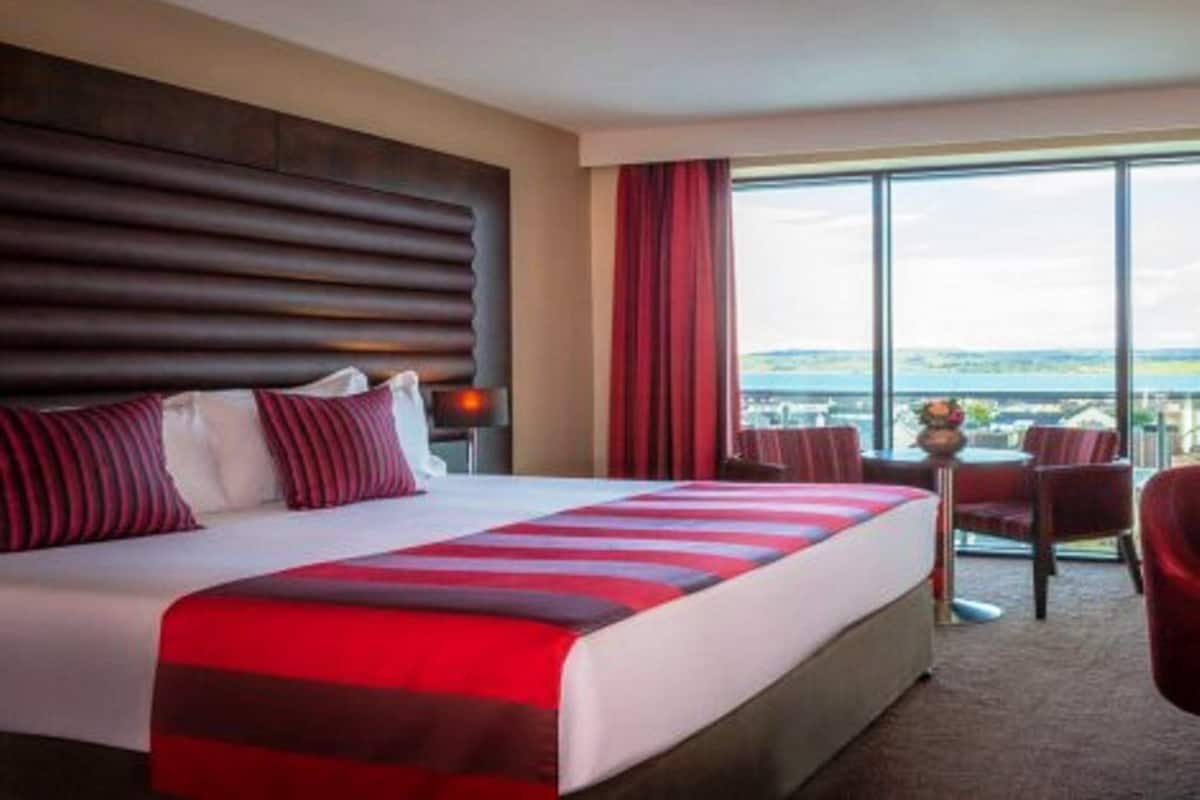 Habitación de hotel con una cama grande, vistas de la ciudad y un balcón con sillas.