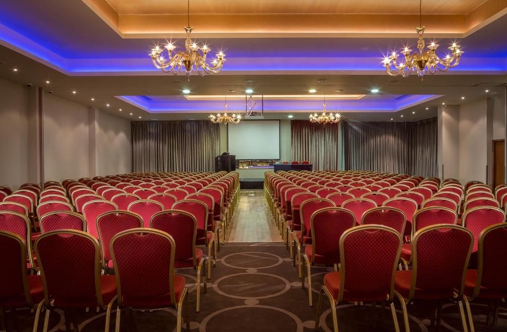 Un salón de conferencias con filas de sillas rojas, un escenario con una pantalla y candelabros.