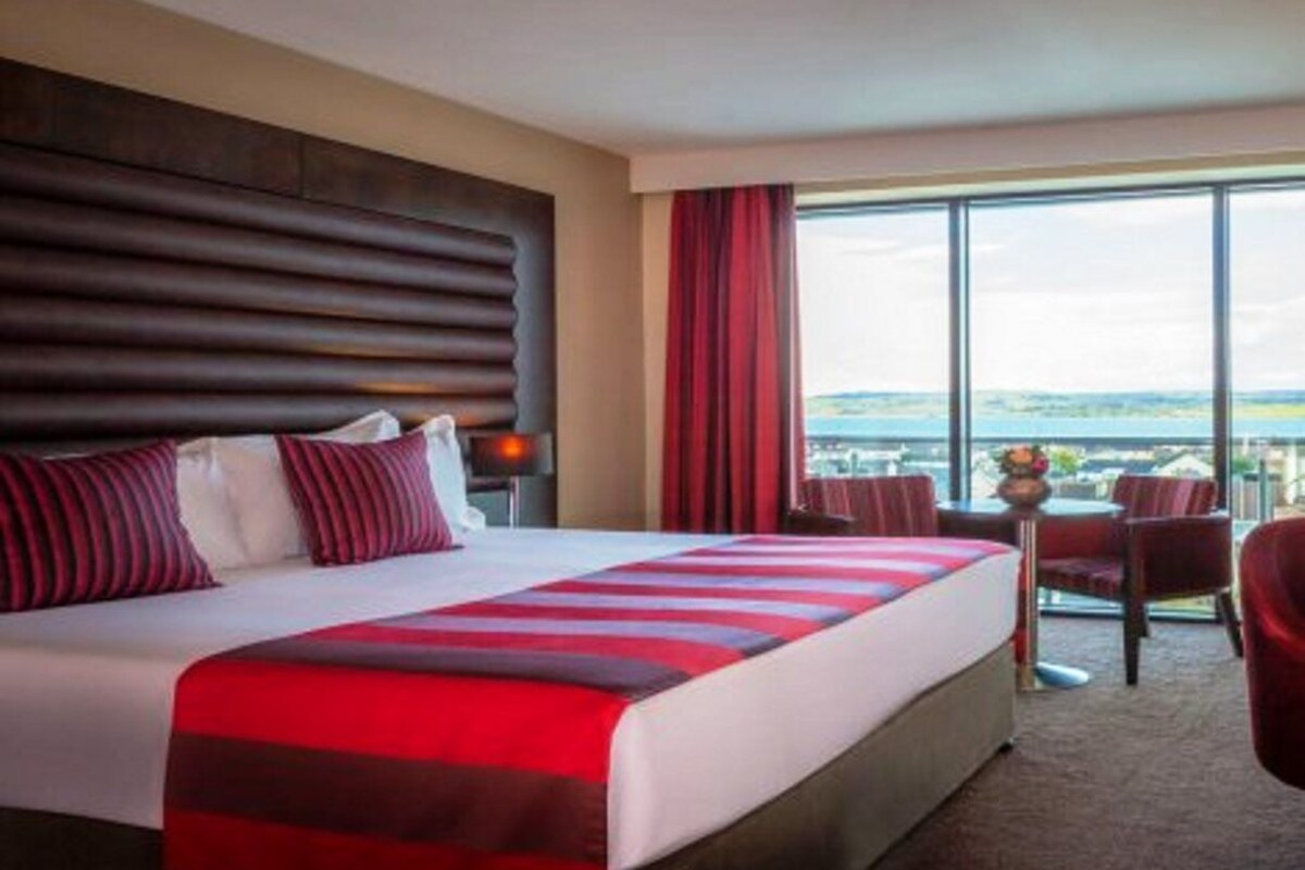 A hotel room with a large bed, a view of the city, and a balcony with chairs.