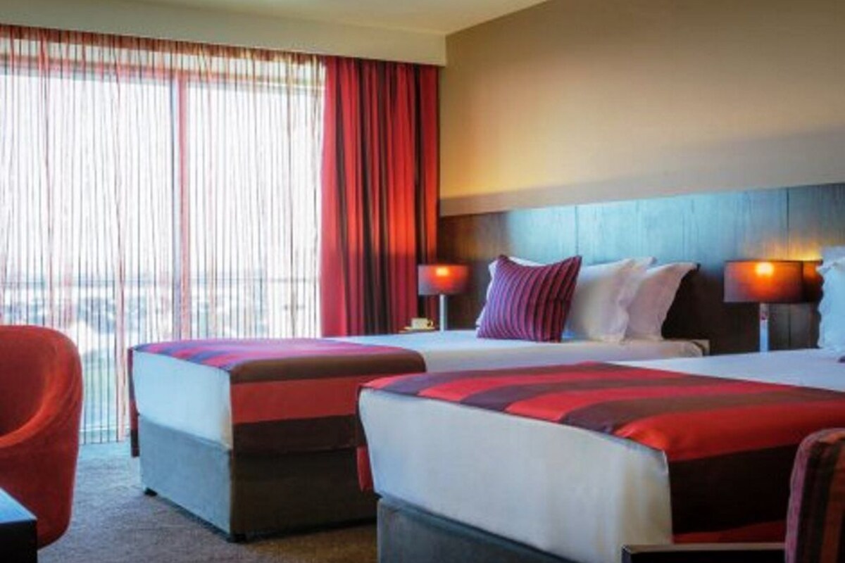 A hotel room with two beds, a red chair, and a view of the city through large windows.