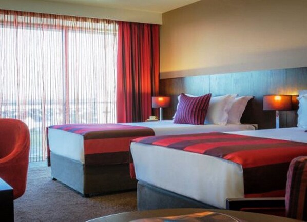 A hotel room with two beds, a red chair, and a view of the city through large windows.