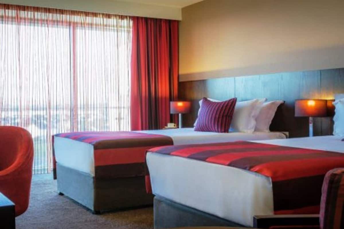 Habitación de hotel con dos camas, una silla roja y vistas a la ciudad a través de amplios ventanales.