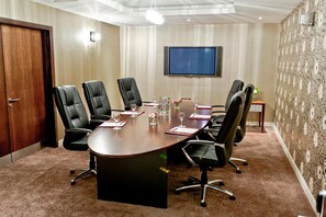 A conference room with a large oval table, six black leather chairs, a flat-screen TV on the wall, and a patterned wallpaper on the right wall.