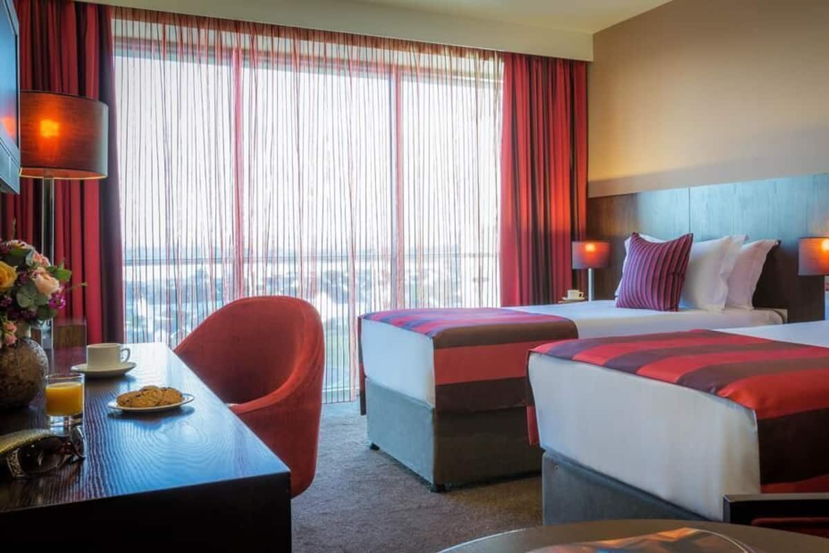 Habitación de hotel con dos camas, un escritorio con silla, una mesita de centro y vistas a la ciudad.