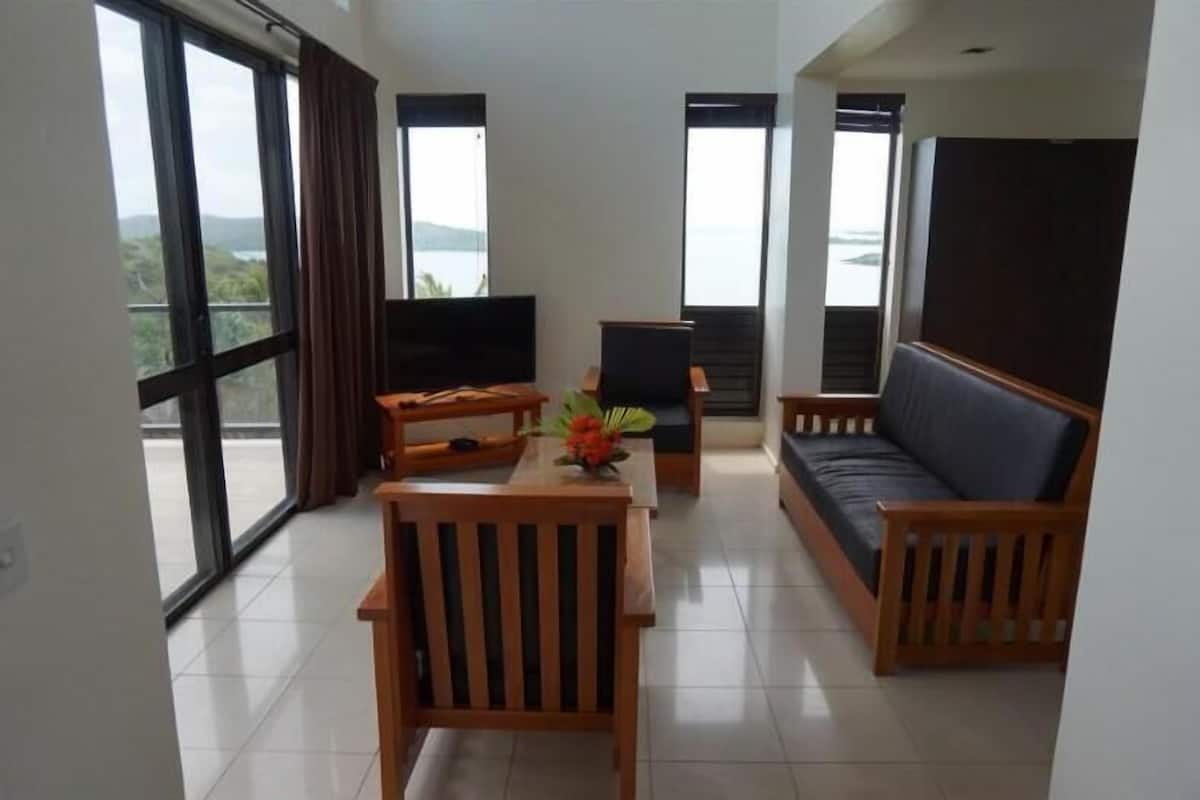 A modern living room with a sofa, wooden chairs, a small table with a flower vase, a TV, and large windows with curtains.