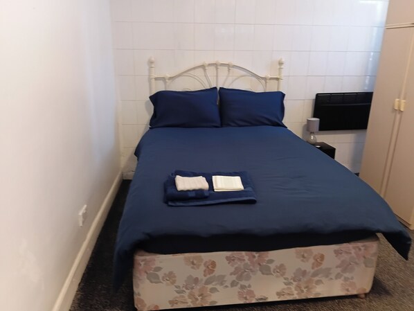 Un lit simple avec une tête de lit, une couvre-lit bleue, deux oreillers et une petite table de chevet avec une lampe.