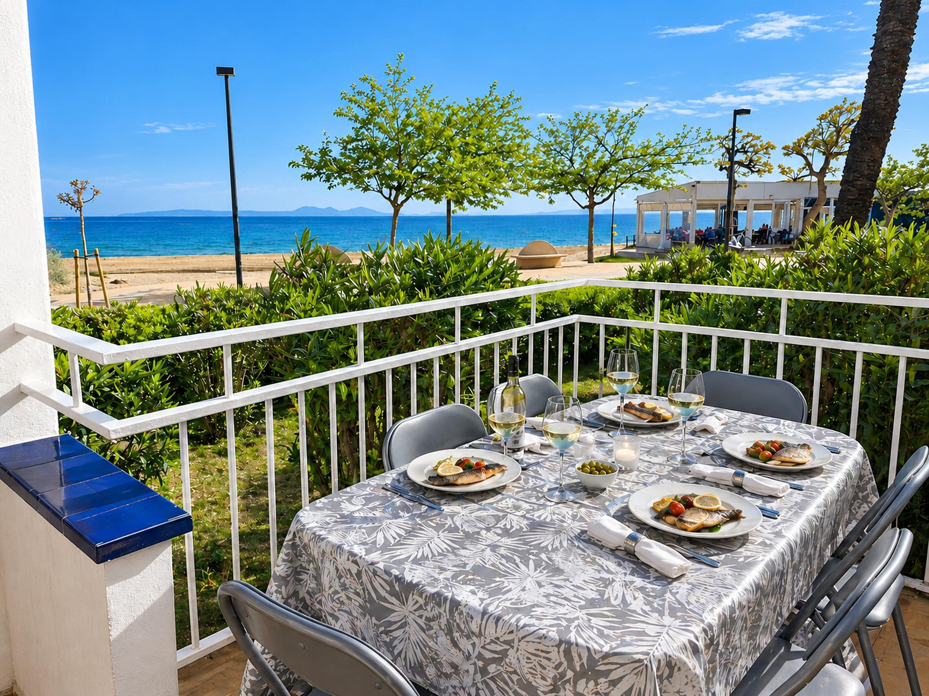 A table set for dining with a view of the sea, a white railing, and greenery.