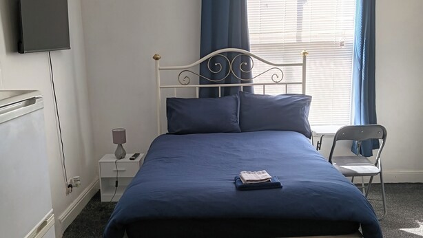 Une chambre bien rangée, avec un lit, un téléviseur fixé au mur, une table de chevet avec une lampe et une chaise.