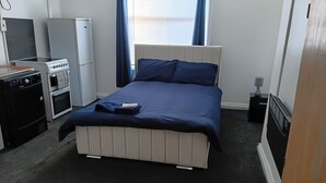 A bedroom with a bed, a small kitchenette, a radiator, and a window with curtains. - Studio Sleeps 2 with Parking (Oldbury)