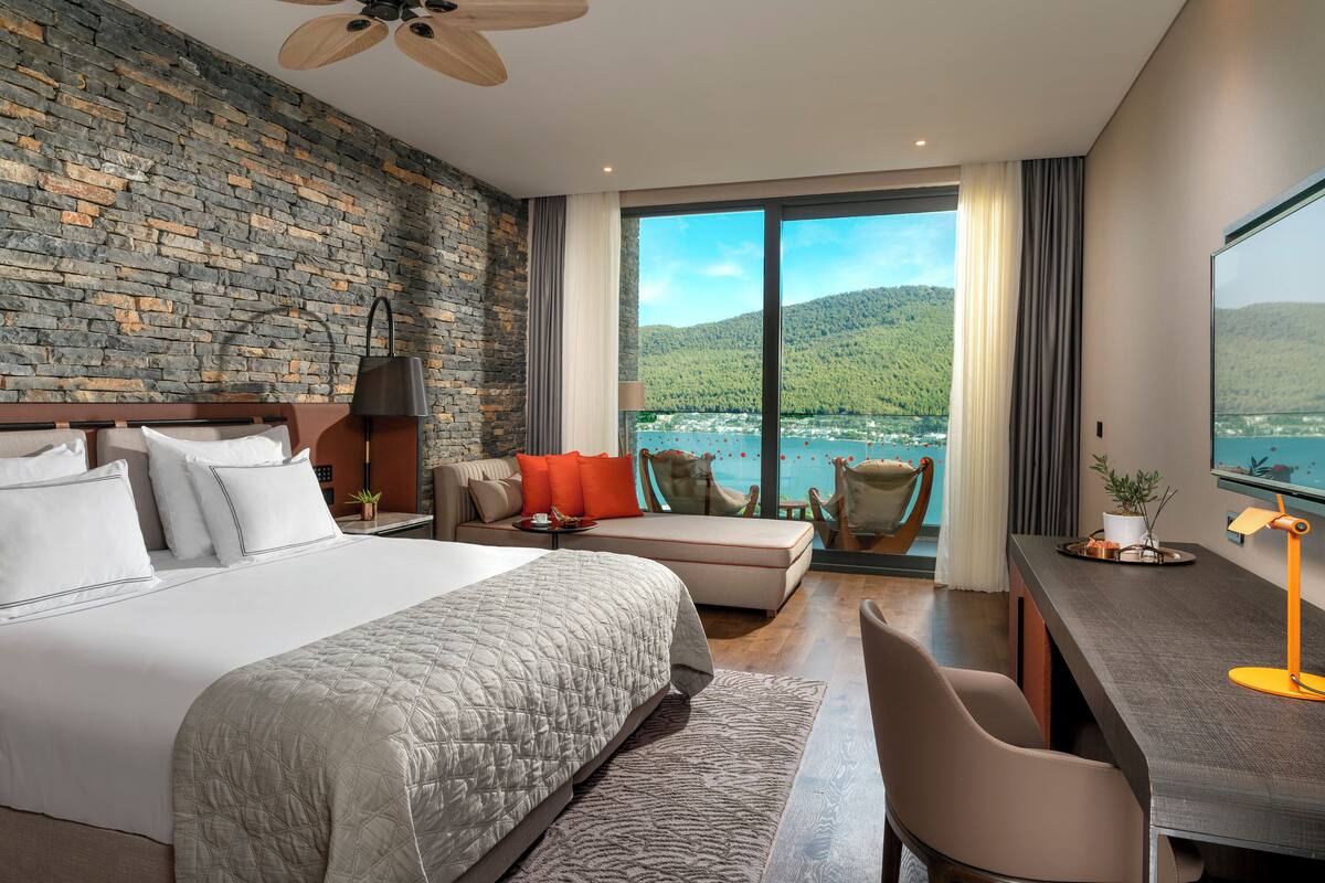 A modern hotel room with a stone wall, a large bed, a sofa, a desk, and a view of a lake and mountains.