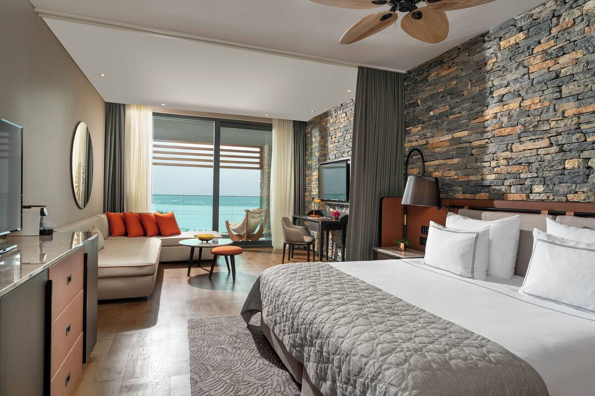 A hotel room with a bed, a desk, a sofa, a TV, and a view of the ocean.